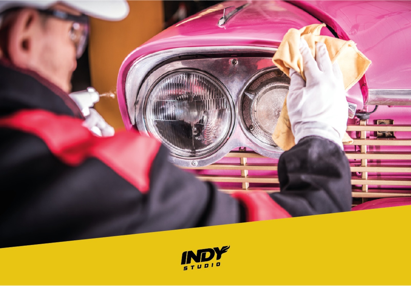 Pessoa com luvas brancas e óculos de proteção limpa o farol cromado de um carro rosa clássico com um pano amarelo.Faixa amarela na base com logotipo Indy Studio.